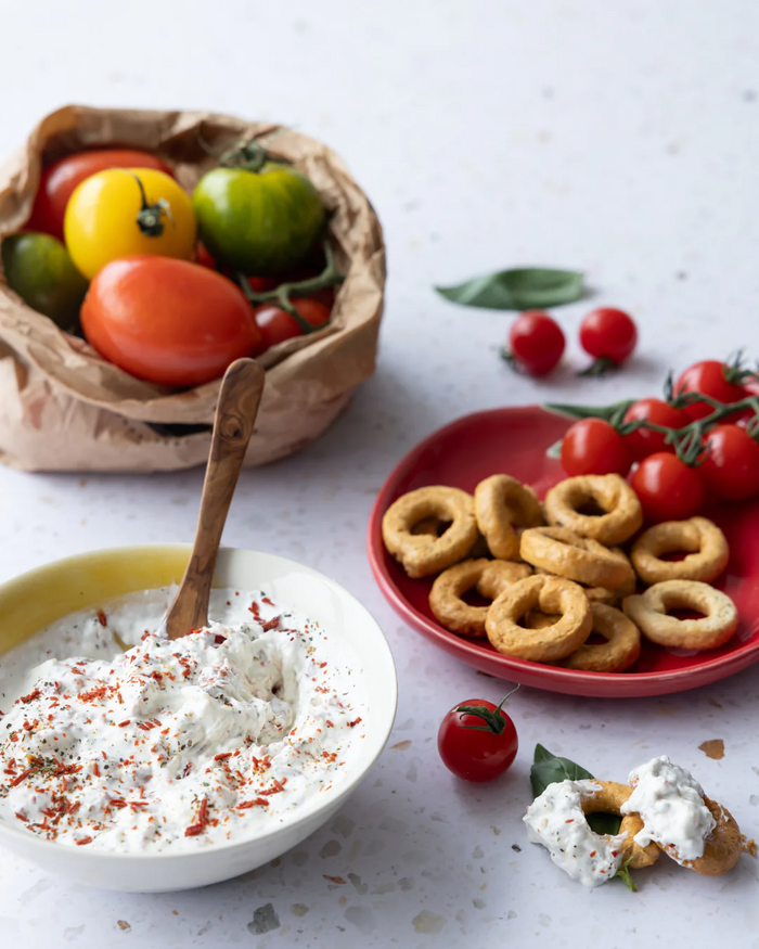 Bowl of burrata dip with herbs on a white counter with tomatoes in a bag behind it and crackers on a red plate behind it