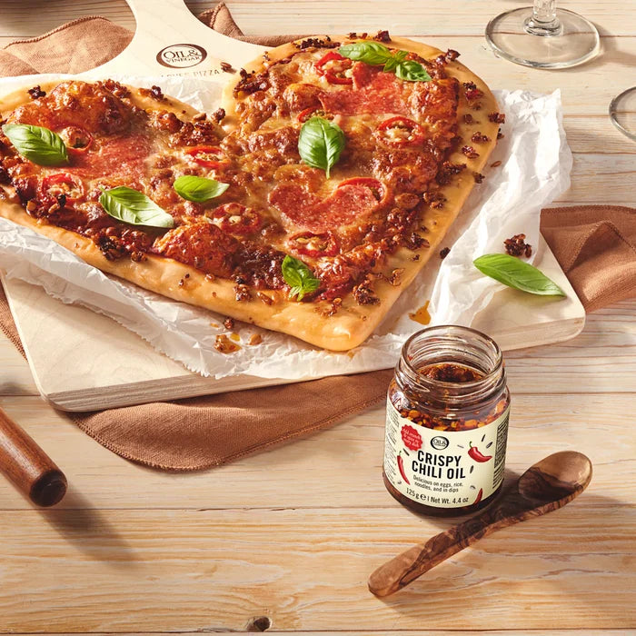 Jar of crispy chili oil on a wooden table with a heart shaped pizza