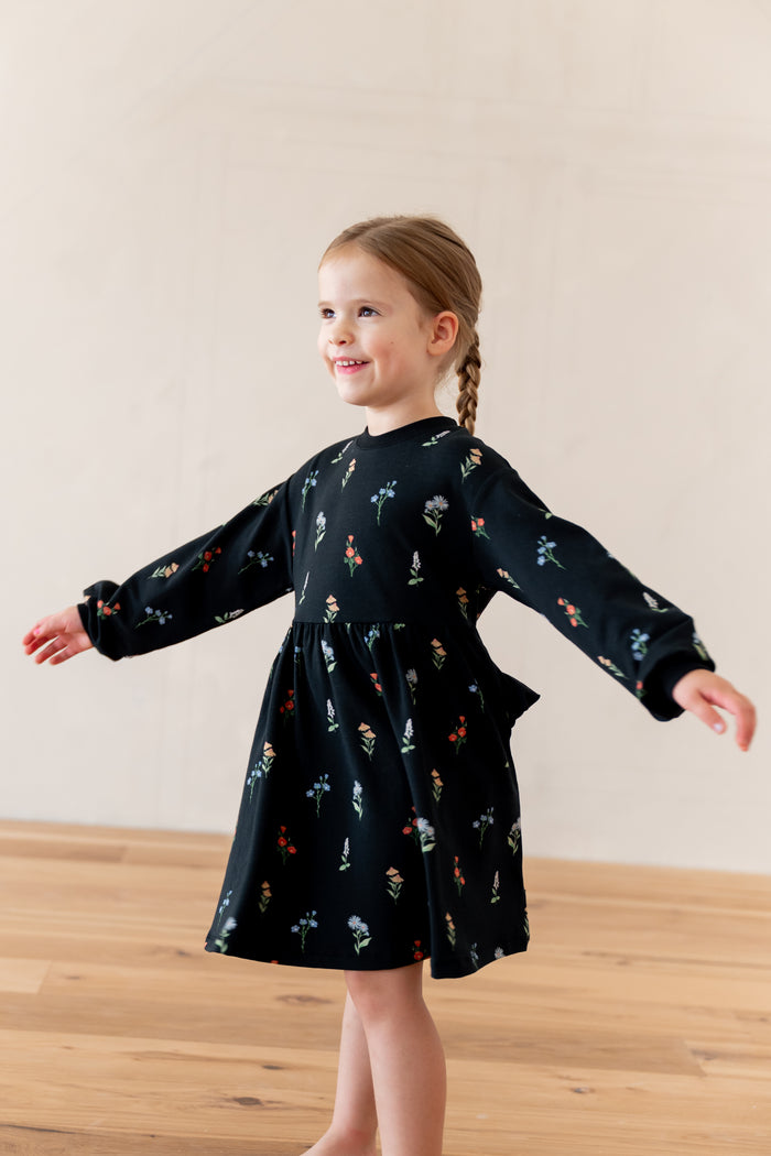 A young girl with a braided ponytail stands in a studio wearing a black long-sleeve dress with a small multicolor floral print. She has her arms outstretched and is smiling, looking to the side.