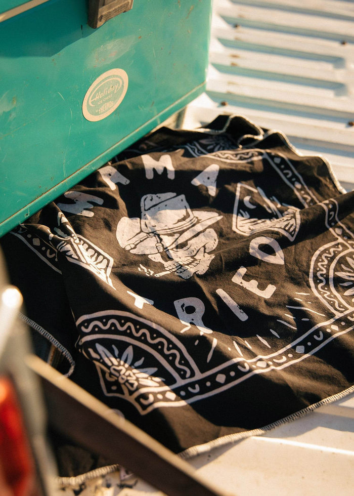 A teal cooler sits on a truck bed beside a black bandana with "Mama Tried" and a cowboy design in white.