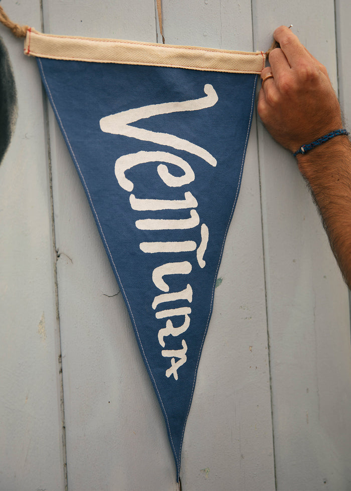 A hand hangs a blue pennant with "Ventura" in white letters on a light wooden wall, evoking a coastal, nautical theme.