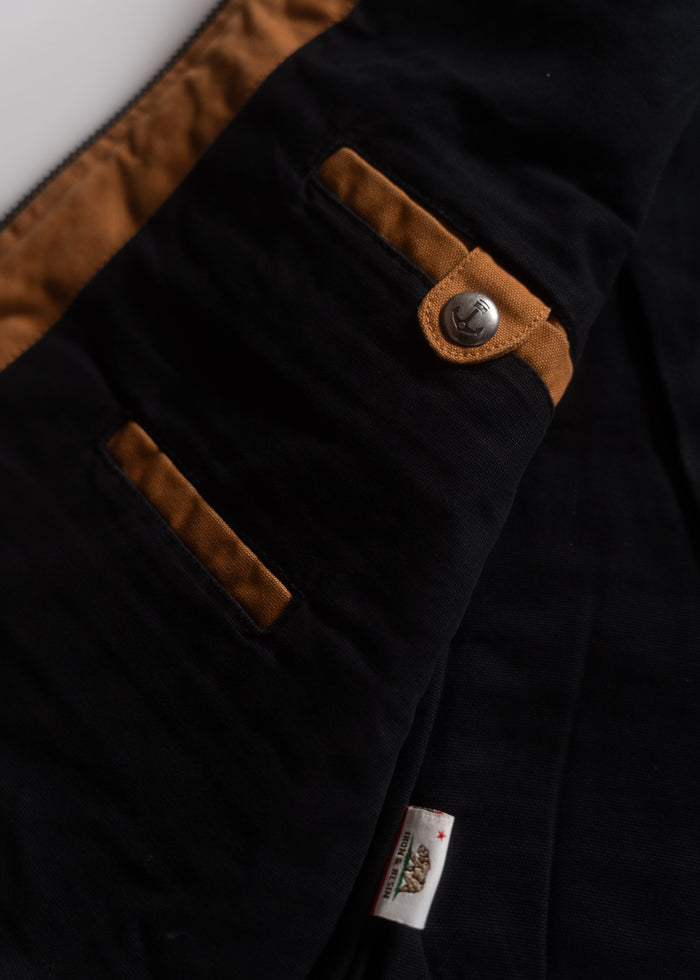 Close-up of a black jacket with brown accents, featuring a button and a visible label. The fabric texture is clearly detailed.