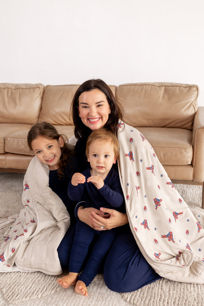 A woman and two kids wearing blue pajamas in front of beige couch wrapped in an oat color Adult Quilted Blanket with Spider-Man print