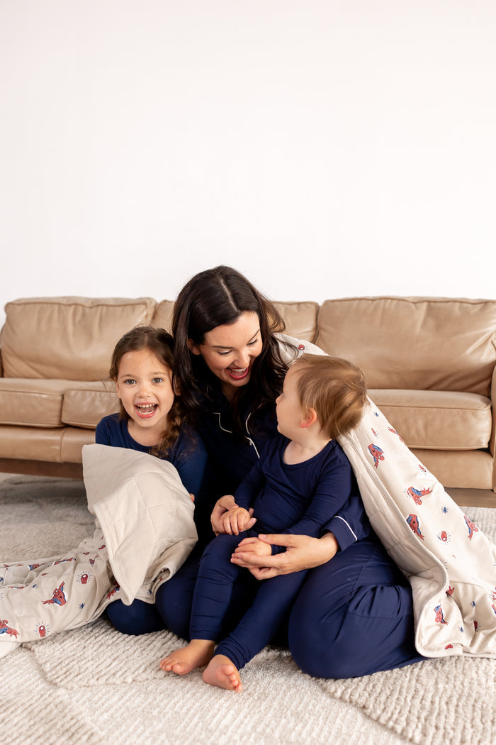 A woman and two kids smiling in blue pajamas in front of beige couch wrapped in an oat color color Adult Quilted Blanket with Spider-Man print