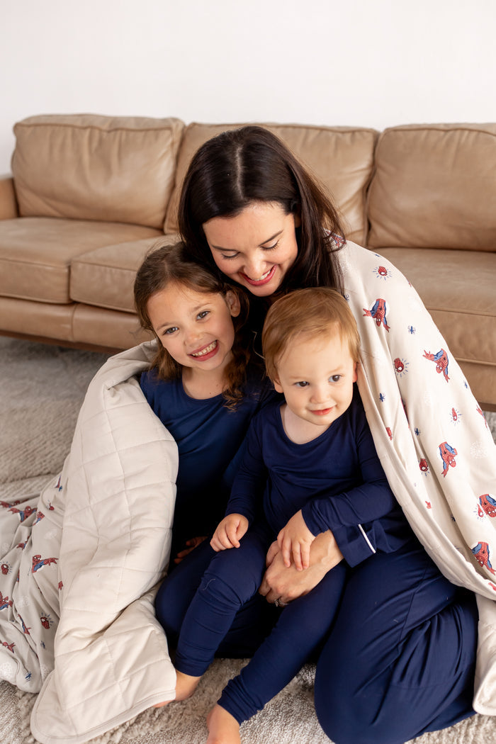 A woman and two kids posing in blue pajamas in front of beige couch wrapped in an oat color Adult Quilted Blanket with Spider-Man print