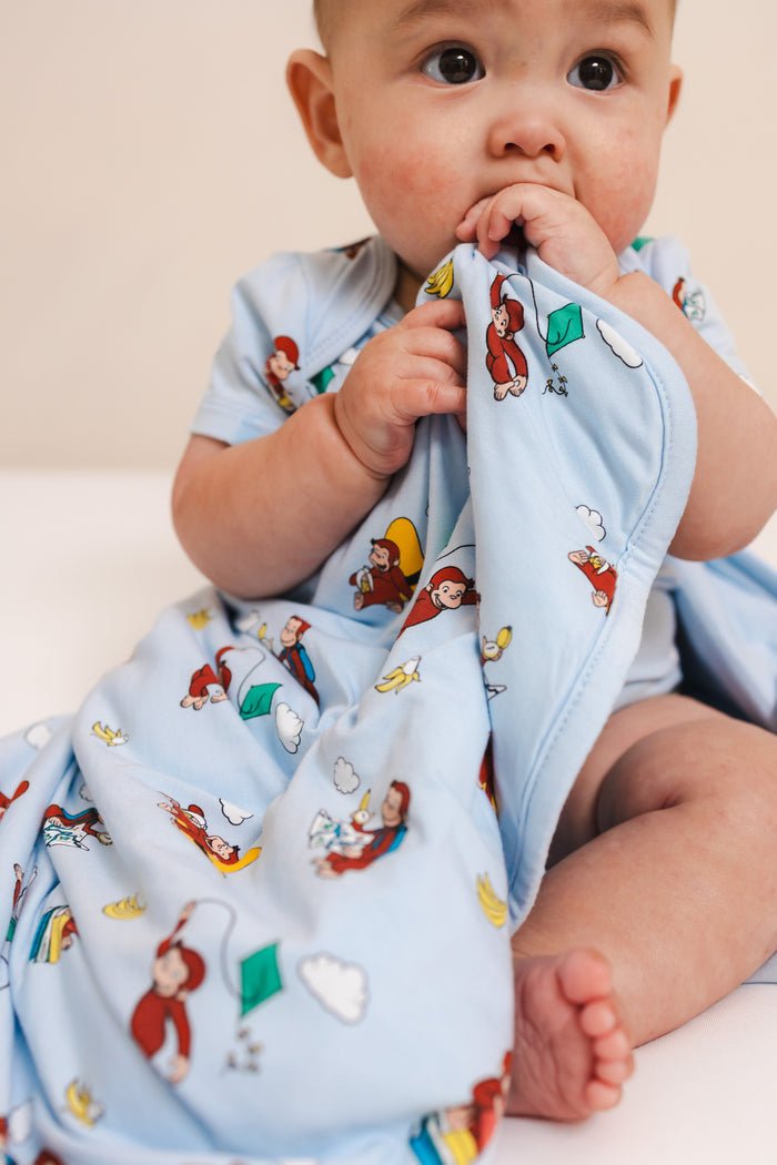 Baby sitting with Baby Blanket in Curious George 1.0 print wearing similar print bodysuit on a beige background