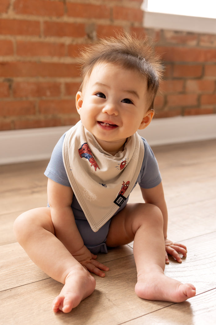 Baby sitting on ground posing in front of a brick wall in an oat color Bib in Spider-Man print over blue bodysuit