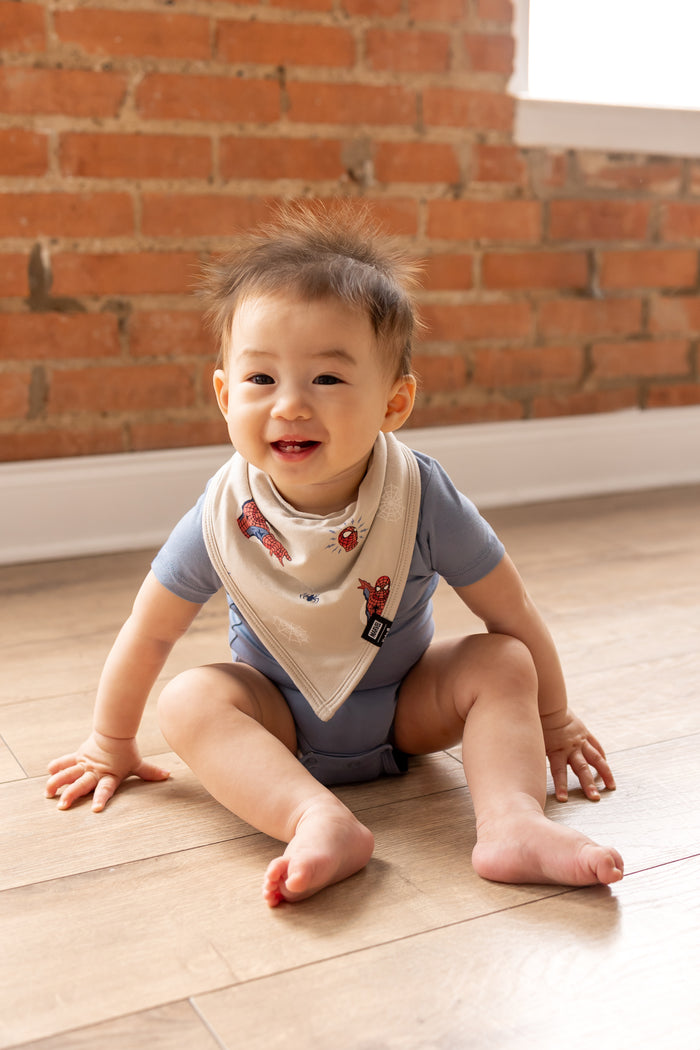 Baby sitting on ground in front of a brick wall wearing an oat color Bib in Spider-Man print over blue bodysuit