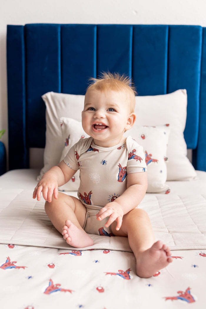 Baby posing on a bed wearing an oat color Bodysuit in Spider- Man print