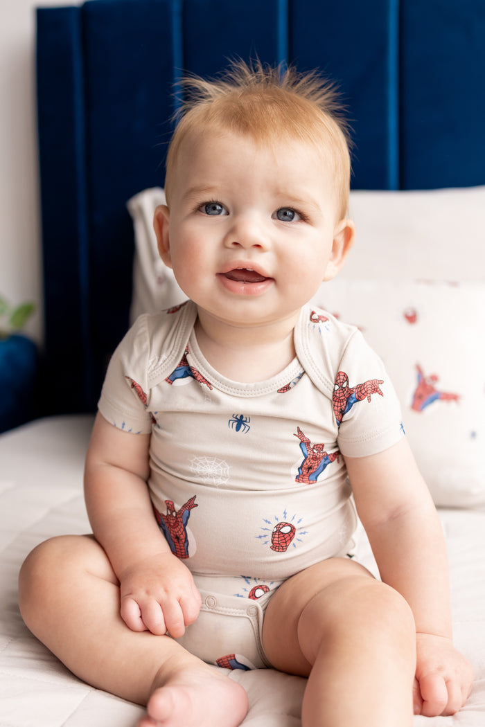 Baby sitting on a bed wearing an oat color Bodysuit in Spider- Man print