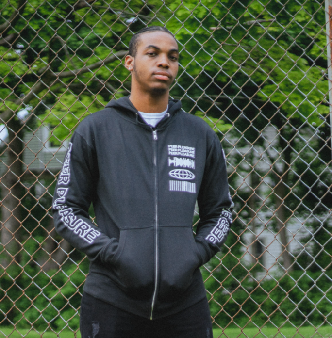 Front of black full zip hoodie. PLEASURE written vertically on one sleeve in white lettering. On the side of the hood , there is a skull drawn in white. On the top corner of the chest, there is a soundwave, globe, and barcode, and text that says PEERPLEASURE drawn in white. An individual is modeling the shirt standing in front of a fence and trees in the back.