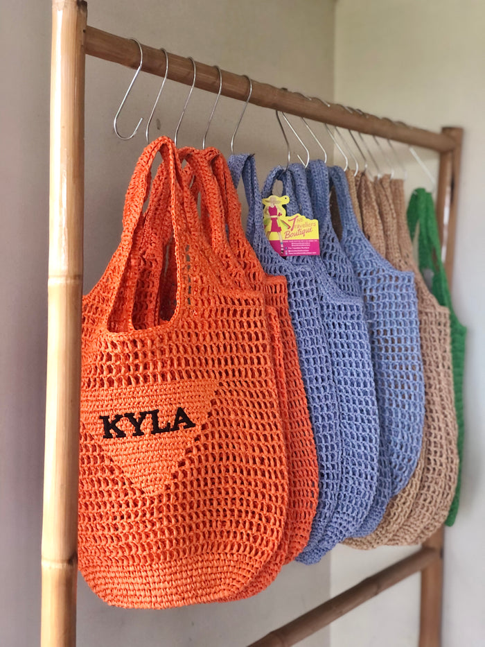 Handmade raffia bucket bag in orange, blue, beige, and green hanging on a wooden rack by Travellers Hat Boutique