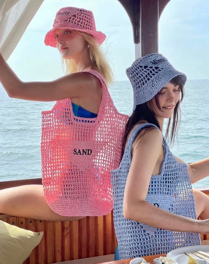 Two women wearing handmade raffia bucket bags in pink and blue while sitting on a boat by the sea