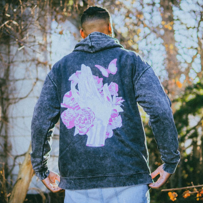 Someone wearing the back of Greyish Black hoodie with a purple hand surrounded by butterflies and flowers which are also purple.