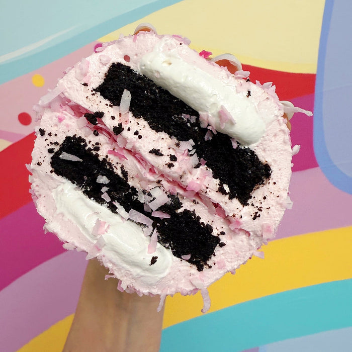 A hand holding a cross section of a XO marshmallow bonbon held against a rainbow background, with layers of coconut marshmallow, extra marshmallow fluff squeezing out and gluten free brownie visible.