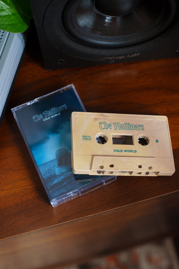 The Flatliners: Cold World Cream Cassette Tape sits on a wooden surface beside its blue-toned case with ocean-themed art, making it a unique addition to any music collection. A speaker is in the background.