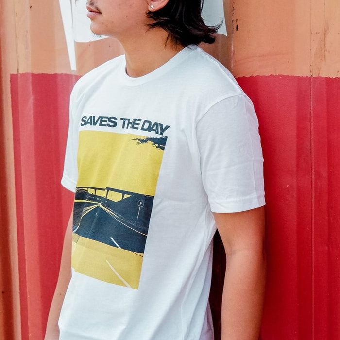 White short sleeve shirt with a yellow tinted image of a highway. There is an overpass and a cloud in the top corner. Above the image is black text that says SAVES THE DAY. An individual is modeling the shirt in front of an orange and red designed wall.