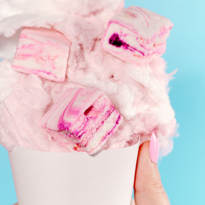 A hand holding a cone full of pink cotton candy with 3 XO Marshmallow gourmet cotton candy marshmallows scattered in it against a blue background