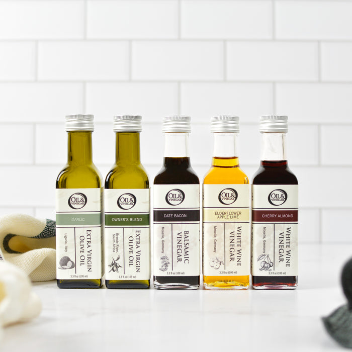 A selection of five oil and vinegar bottles in 100mL bottles, displayed in a row on a kitchen countertop, with decorative elements in the background.