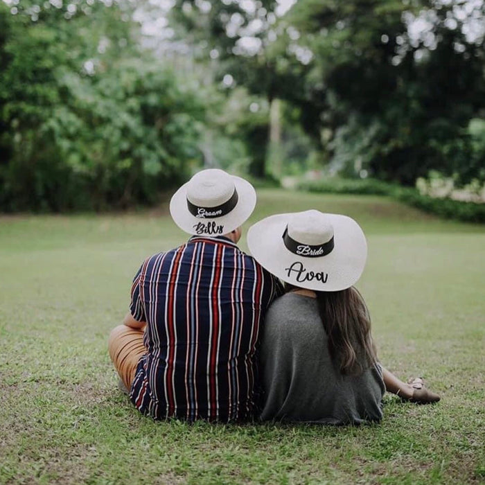 Couple sitting on grass wearing custom prenuptial hat set with personalized names from Travellers Hat Boutique