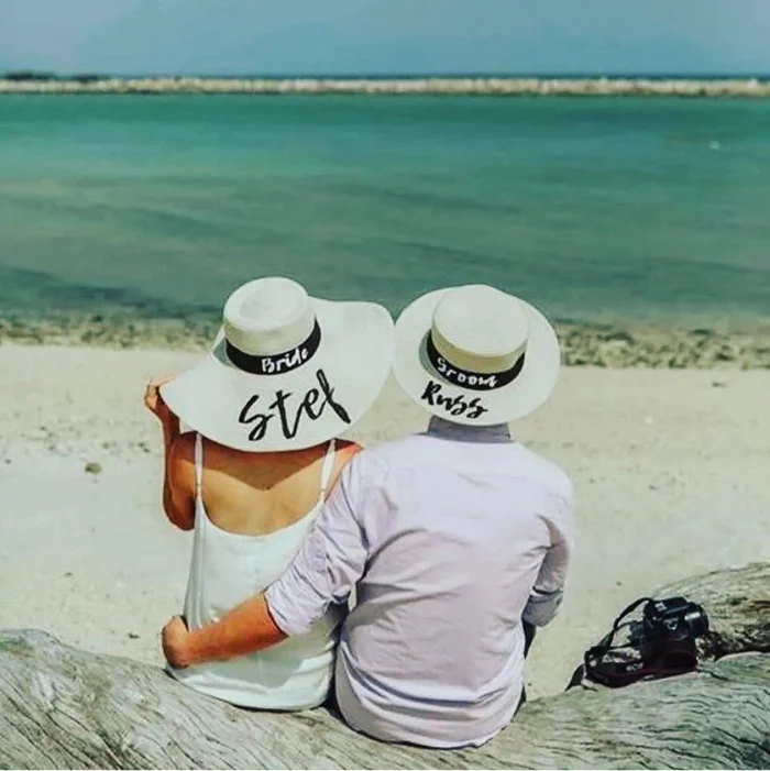 Bride and groom wearing custom prenuptial hat set with names on a beach by the ocean