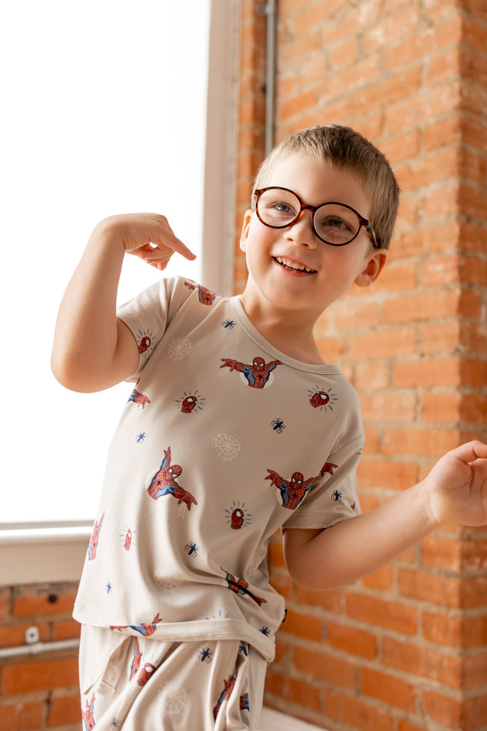 Boy with specs posing in oat color Toddler Basic Tee and Toddler Drawstring shorts with Spider-Man print in front of a brick wall
