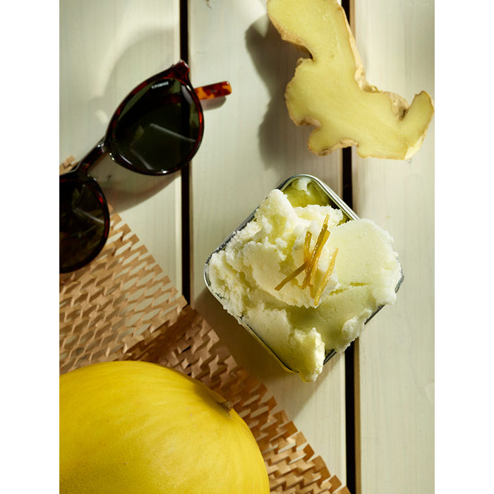 Das Unold Eisbuch – 101 Eisrezepte zeigt einen Metallbehälter mit hellgrünem Eis mit Ingwerstreifen, der auf einem weißen Holztisch neben frischem Ingwer, gelber Melone und einer Sonnenbrille steht.
