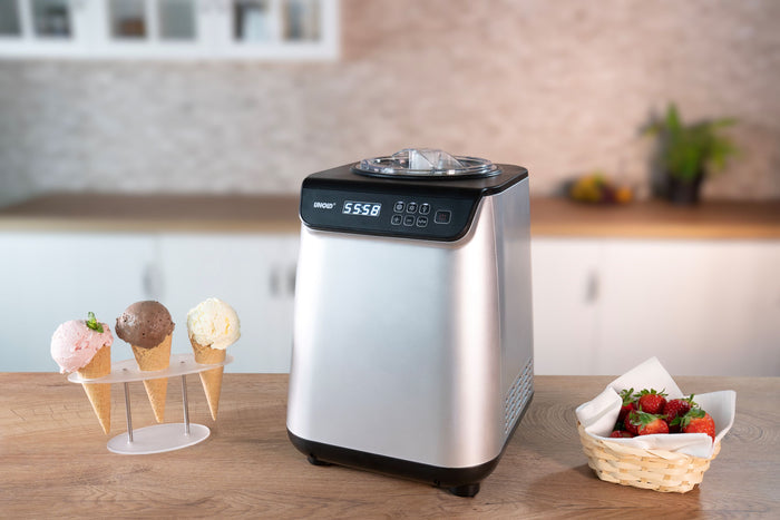 Die Unold Eismaschine Uno 1,2 L steht auf einer hölzernen Küchentheke neben einem Ständer mit drei Eistüten und einem Korb Erdbeeren. Ihre Digitalanzeige zeigt 55:58.