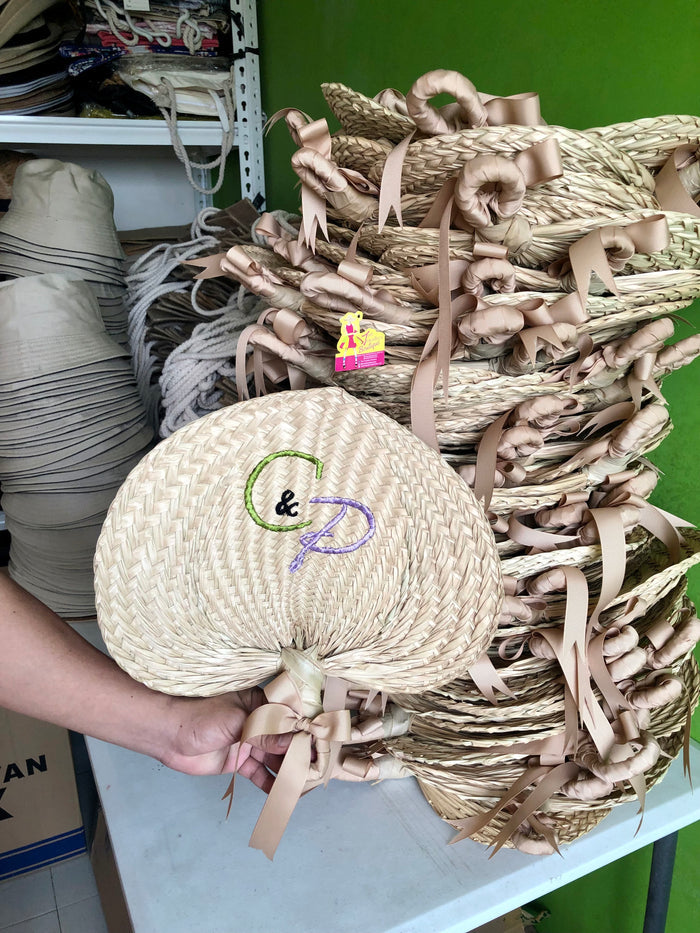 Hand holding a personalized embroidered abaniko fan with ribbons in front of stacked woven fans