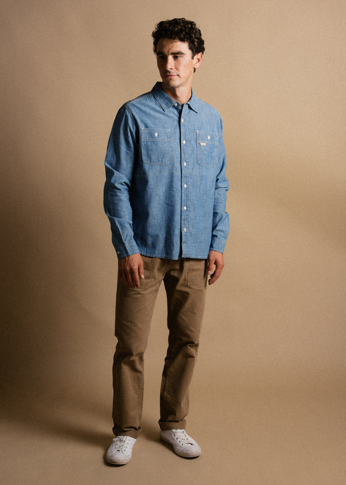 Man in blue denim shirt and khaki pants stands against a beige background, wearing white sneakers, looking to the side.