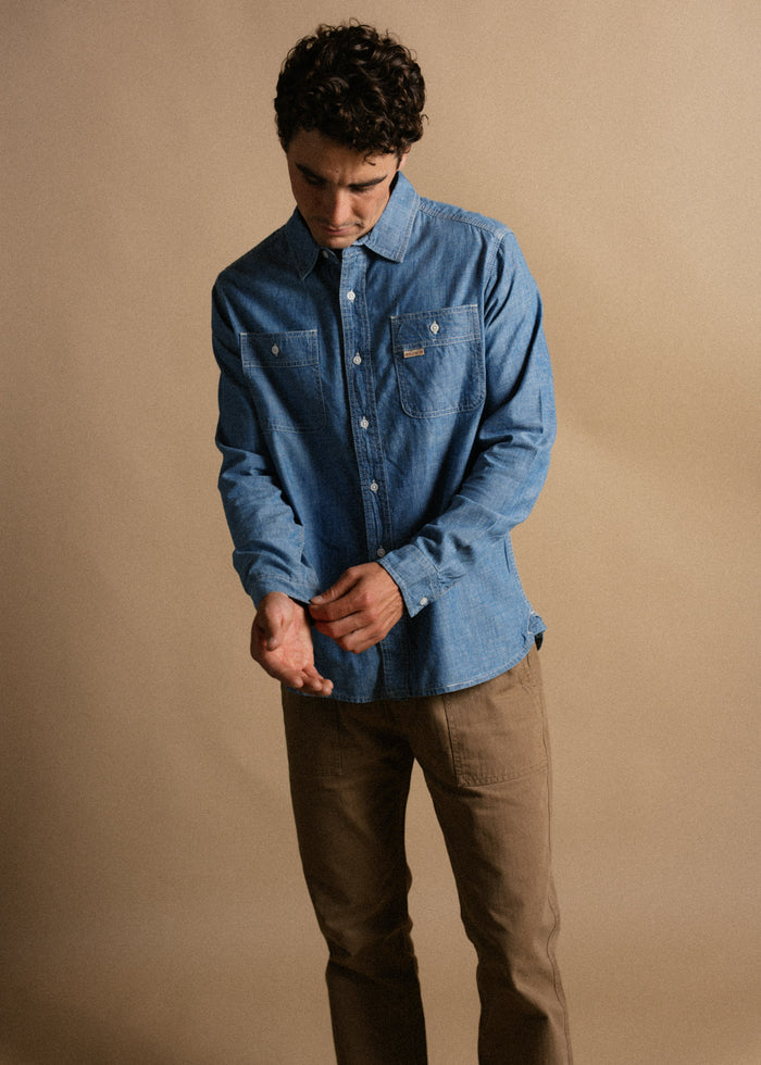 Man in a blue denim shirt adjusts sleeve, wearing brown pants, against a beige background, looking down thoughtfully.