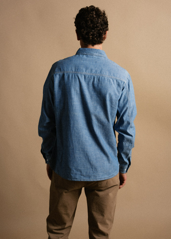 Man in blue denim shirt and khaki pants stands with back to camera against a beige background, showcasing casual fashion style.