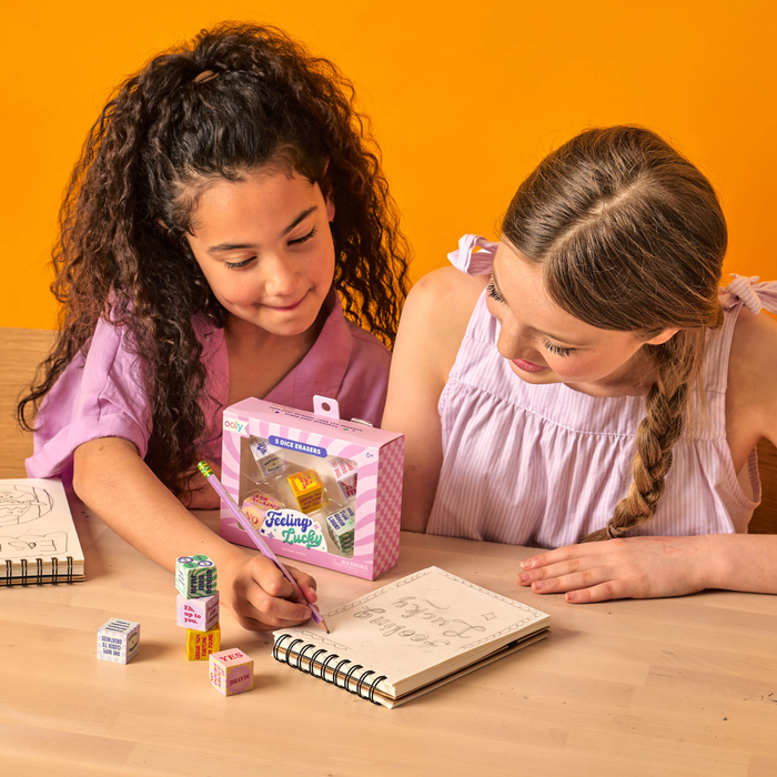Two girls drawing in a sketchbook with OOLY Feeling Lucky Dice Erasers at their side on a wooden table