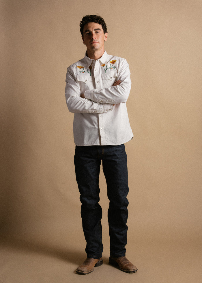Man in embroidered white shirt, jeans, and boots stands confidently against a beige background, arms crossed.
