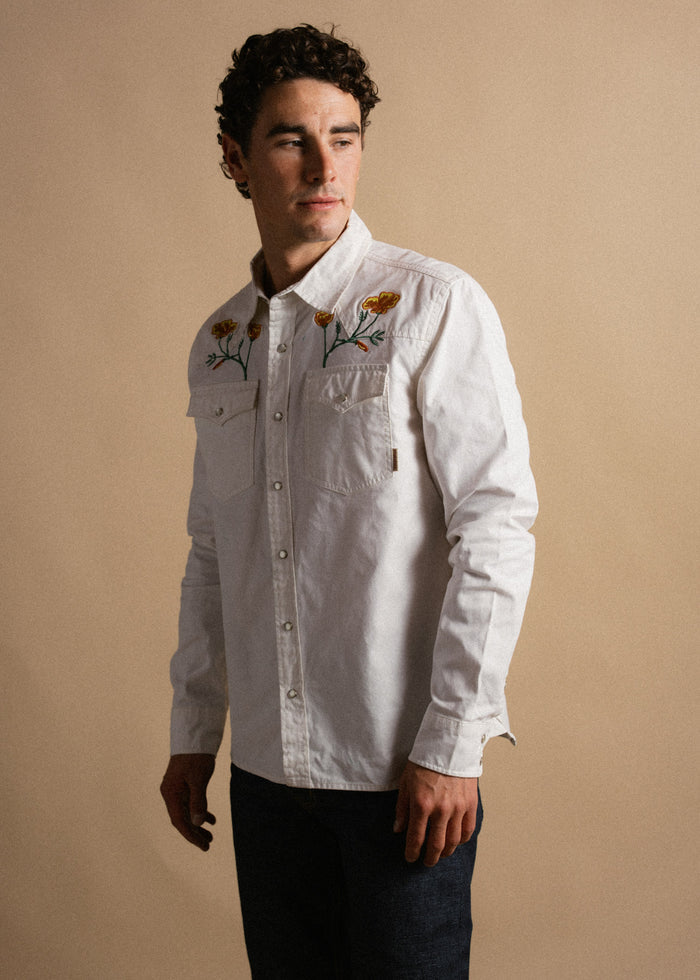 Man in a white embroidered shirt with floral patterns, looking to the side against a beige background.