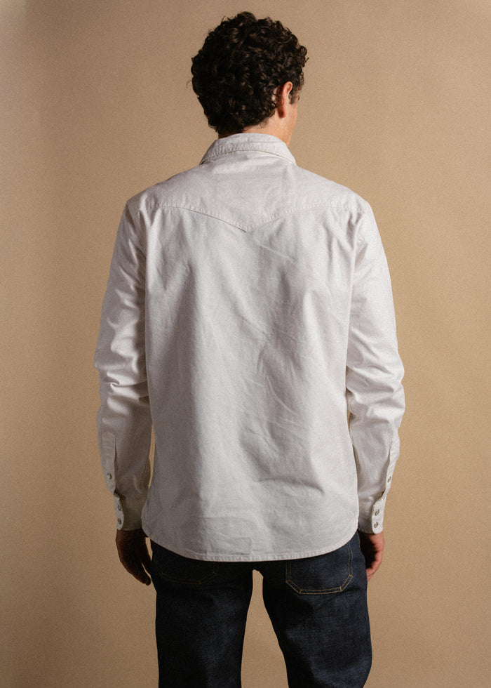 Man with curly hair in a white shirt and jeans, seen from behind, standing against a beige background.