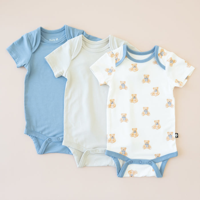 Three baby bodysuits in blue, off-white, and patterned with teddy bears on a beige background.