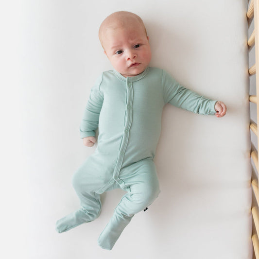 Newborn laying in crib and wearing Kyte Baby Footie in Sage