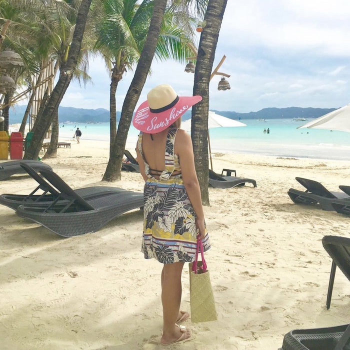 Woman wearing a hand-painted straw hat with pink brim on a tropical beach with palm trees and lounge chairs