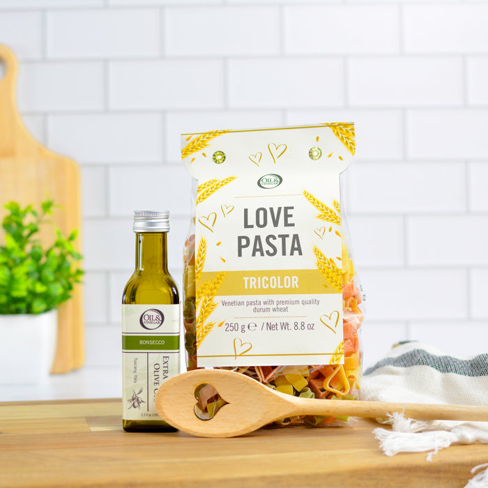 Bag of heart shaped pasta, 100 ml bottle of olive oil and a wooden spoon on a wooden board with a white background