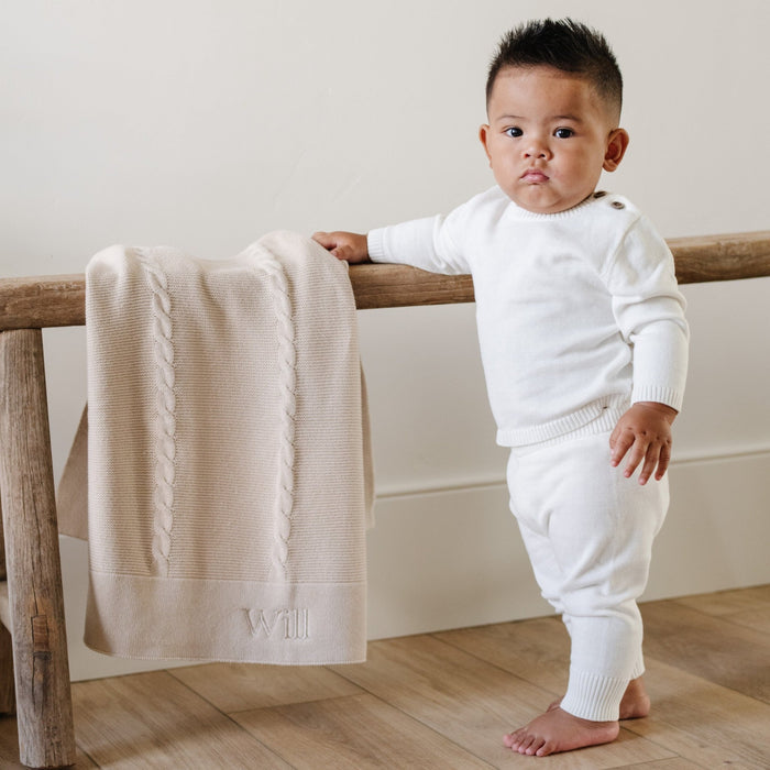 A baby boy stands stands by a Saranoni Heirloom Knit Blanket. He is wearing an all-white, 100% cotton baby outfit that combines style and comfort. The Saranoni cloud-soft cotton set features a breathable waistband, loosely fitted sleeves, and elevated button details, making it perfect for everyday wear or special occasions. This heirloom-quality baby clothing offers timeless style and ultimate comfort. Ideal for parents searching for luxury baby clothes, soft cotton baby sets, or high-quality in