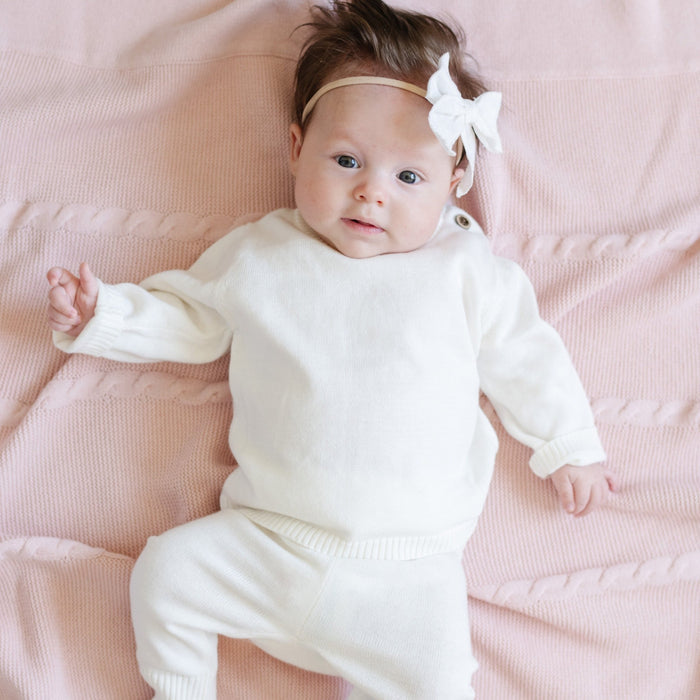 A baby girl lays on a Saranoni Heirloom Knit Blanket. She is wearing an all-white, 100% cotton baby outfit that combines style and comfort. The Saranoni cloud-soft cotton set features a breathable waistband, loosely fitted sleeves, and elevated button details, making it perfect for everyday wear or special occasions. This heirloom-quality baby clothing offers timeless style and ultimate comfort. Ideal for parents searching for luxury baby clothes, soft cotton baby sets, or high-quality infant ou