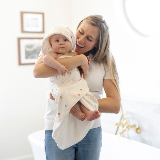 Smiling mom holds her baby wrapped in a Saranoni luxury hooded towel, featuring an ultra-soft fabric for quick-drying comfort. This plush baby towel is perfect for bath time, pool time, or beach days, offering a cozy hood and gentle fabric designed for newborns, infants, and toddlers. Adorable gender-neutral baby towel. A must-have baby bath essential, baby shower gift, and new mom must-have. The hooded towel has an ice cream print. The ultimate newborn hooded towel and baby bath towel.