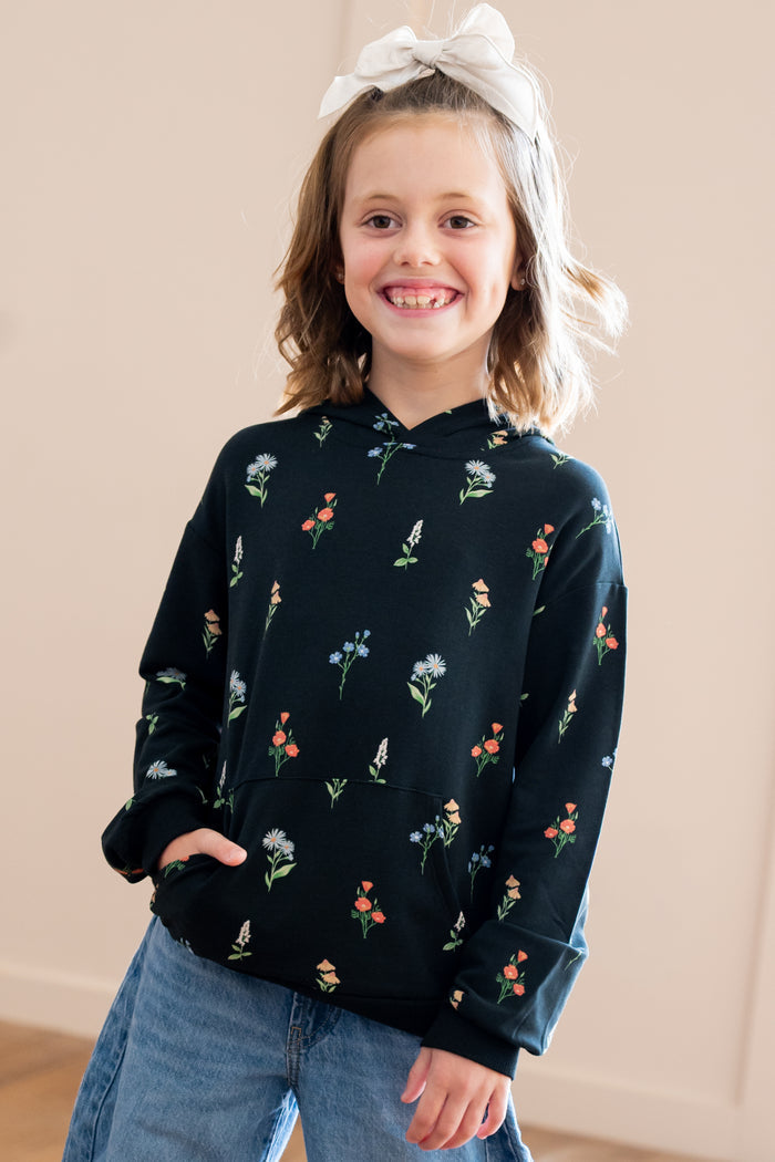 A young girl smiling and wearing a black long-sleeve hoodie with a colorful floral print, paired with blue jeans and a white bow headband, standing against a plain beige wall.