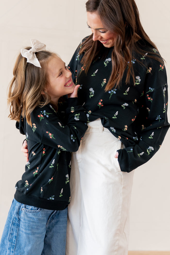 A woman and a young girl wearing matching black long-sleeve sweatshirts with a small multicolor floral print, standing together against a plain beige background.