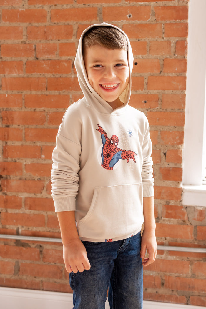 Boy posing with an oat color hoodie cap on his head in front of a brick wall with a Spider-Man print in center over a blue jeans
