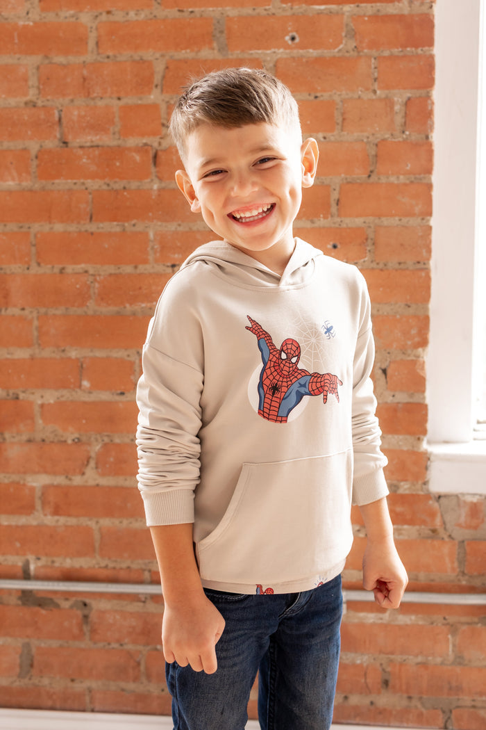 Boy standing in front of a brick wall wearing an oat color Hoodie with a Spider-Man print in center over a blue jeans