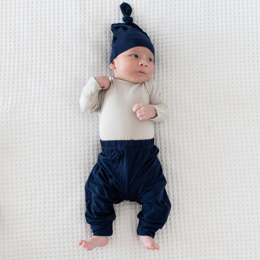 Newborn wearing Kyte Baby Knotted Cap in Navy with matching pants and a Long Sleeve Bodysuit in Oat