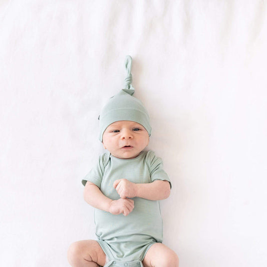 Newborn laying on a crib sheet in Oat while wearing  a Bodysuit and Knotted Cap in Sage