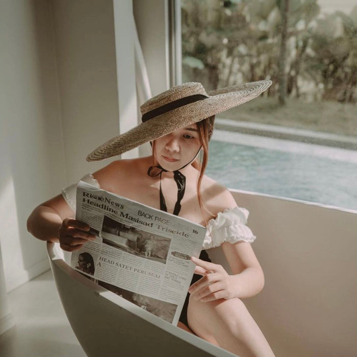 Woman wearing a wide-brimmed straw hat with black ribbon sitting in a bathtub reading a newspaper near a window